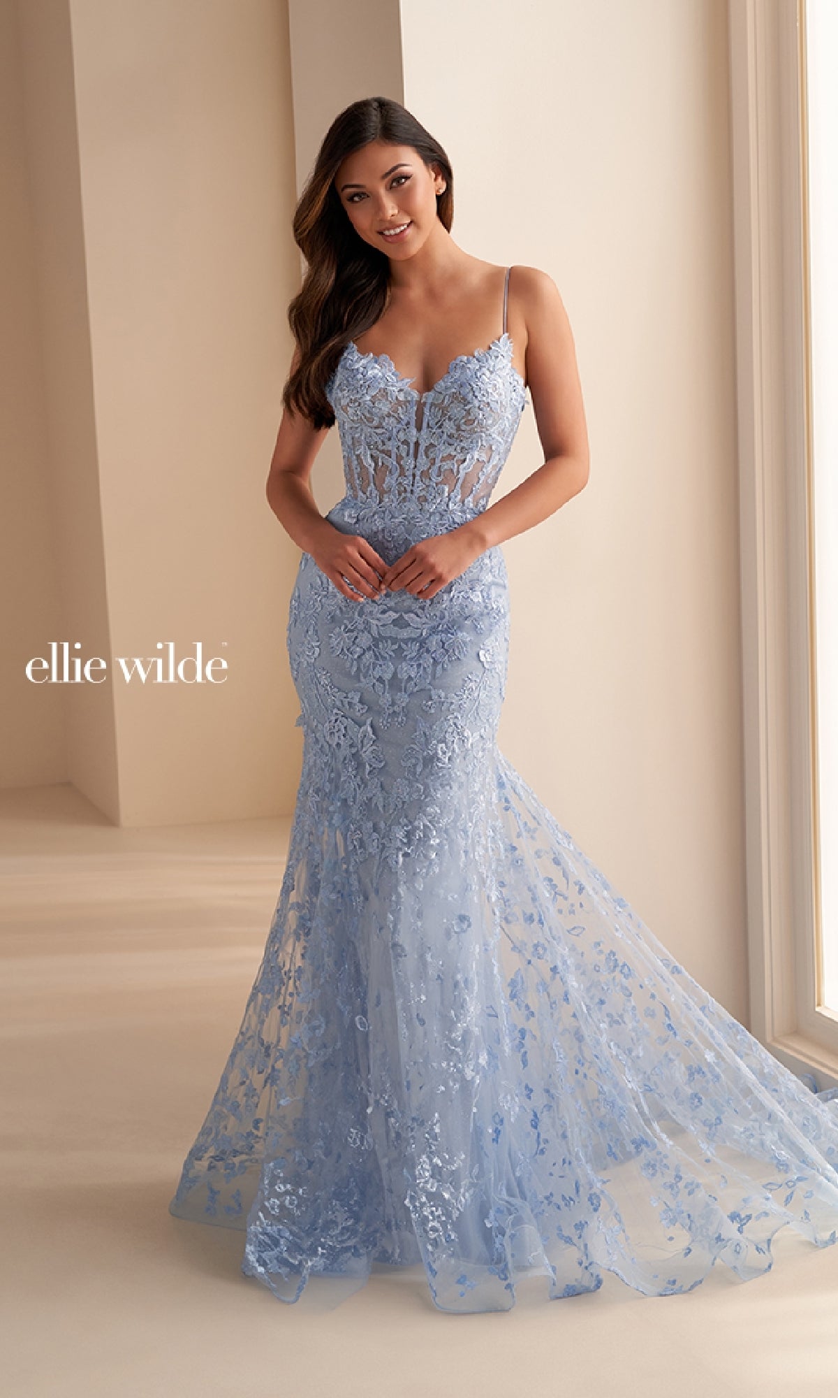 A woman stands indoors by a large window, wearing the Ellie Wilde EW36008 long embroidered prom dress in light blue with lace accents and thin straps.
